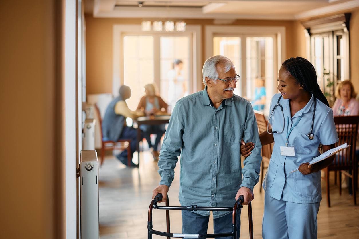 A clinician walking beside a resident using a walker, along a corridor in a skilled nursing facility.